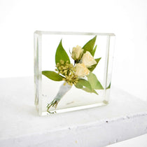A small floral block preservation featuring a boutonniere with white roses and green leaves, set within a clear resin block on a white background.