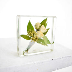 A small floral block preservation featuring a boutonniere with white roses and green leaves, set within a clear resin block on a white background.